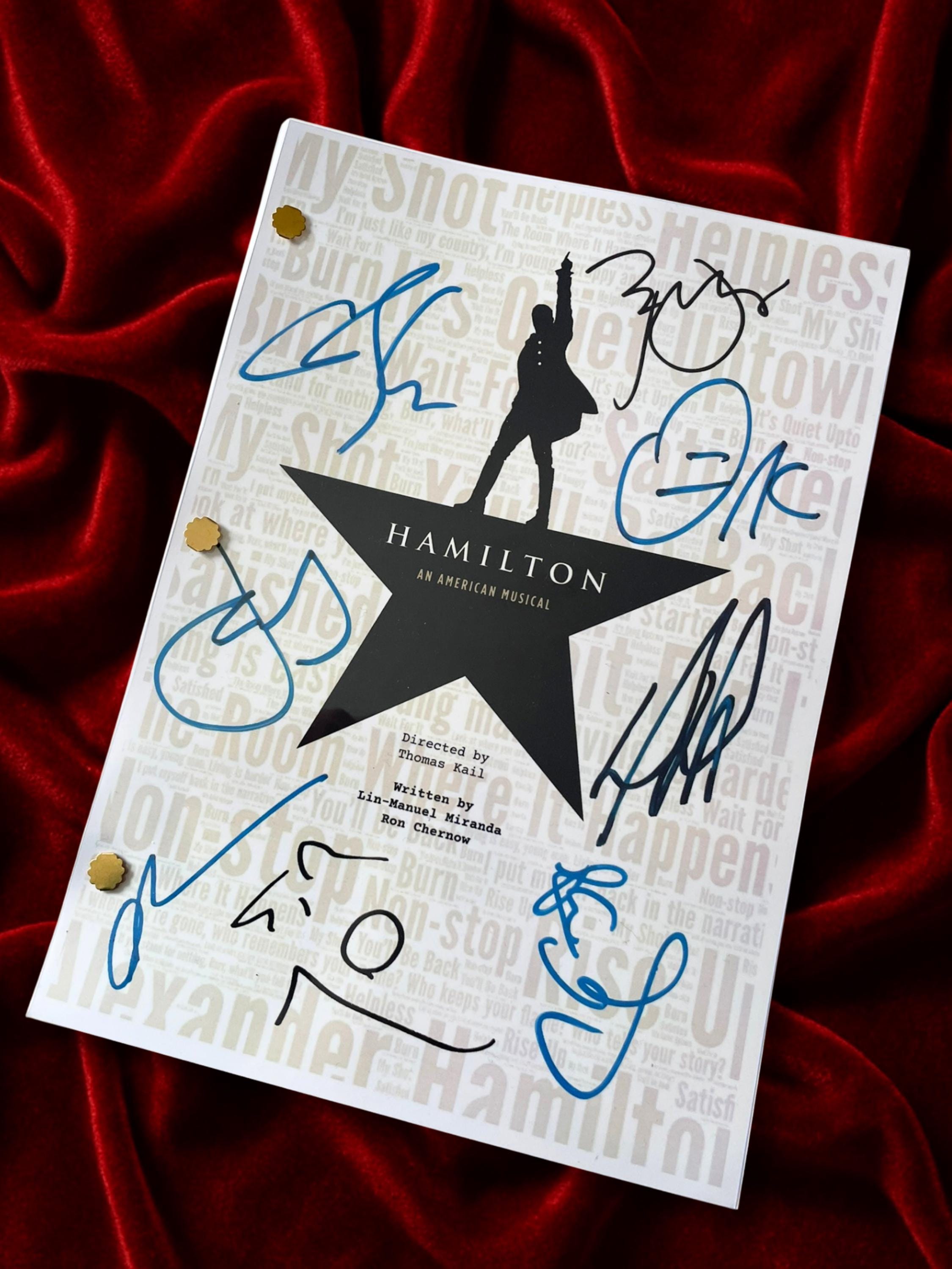 A white script featuring HAMILTON The Musical and a silhouette of Lin-Manuel Miranda as Hamilton on a star, with multiple signatures in blue and black ink. Set against a deep red velvet background.