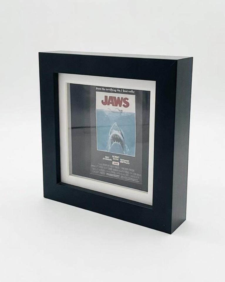 A framed movie poster for the film "Jaws." The poster features a menacing shark with the title "JAWS" in bold letters. The frame is black and has a white mount around the poster.