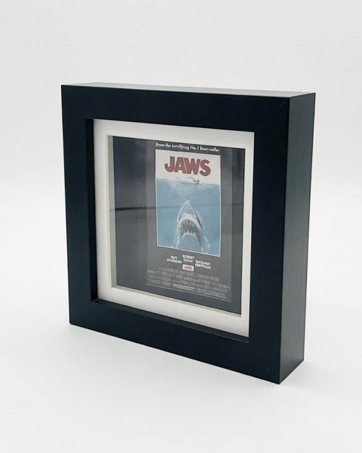 A framed movie poster for the film "Jaws." The poster features a menacing shark with the title "JAWS" in bold letters. The frame is black and has a white mount around the poster.
